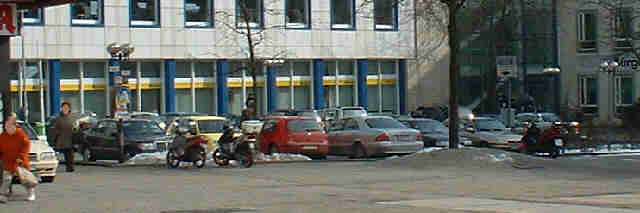 Foto: Motorräder im Vordergrund, dahinter ein Parkplatz. Hinter dem Parkplatz das Postgebäude.